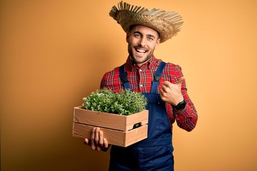 Young rural farmer man wearing countryside hat holding green organic plant from harvest happy with big smile doing ok sign, thumb up with fingers, excellent sign