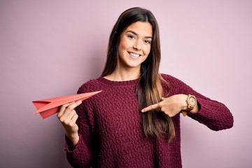 Sticker - Young beautiful girl holding paper plane standing over isolated pink background with surprise face pointing finger to himself