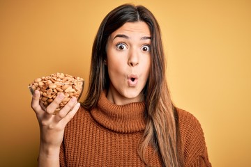 Sticker - Young beautiful girl holding bowl of salty peanuts standing over yellow background scared in shock with a surprise face, afraid and excited with fear expression