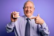 Grey haired senior man holding orthodontic prosthesis denture over purple background with surprise face pointing finger to himself