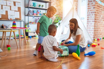 Poster - Young beautiful teacher and toddlers playing around lots of toys at kindergarten