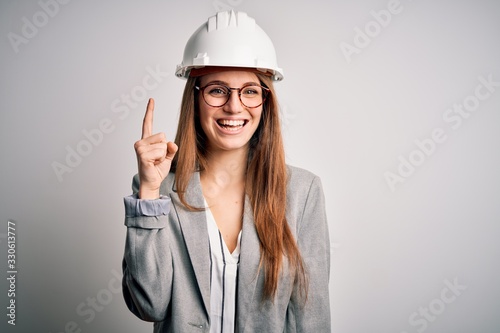 Fotografía Young beautiful redhead architect woman wearing security helmet over white background pointing finger up with successful idea