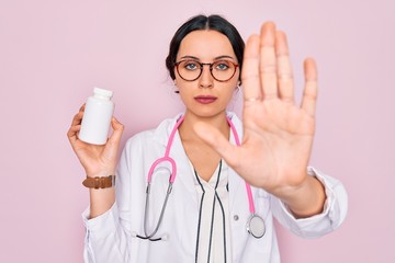 Wall Mural - Young beautiful doctor woman with blue eyes wearing stethoscope holding medicine pills with open hand doing stop sign with serious and confident expression, defense gesture