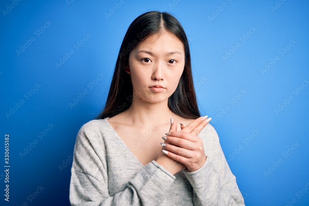 Young beautiful asian woman wearing casual sweater standing over blue isolated background Holding symbolic gun with hand gesture, playing killing shooting weapons, angry face