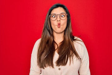 Sticker - Young hispanic smart woman wearing glasses standing over red isolated background making fish face with lips, crazy and comical gesture. Funny expression.