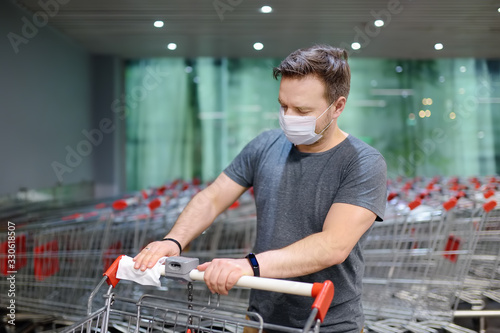 Cuadros en Lienzo Man wearing disposable medical face mask wipes the shopping cart handle with a disinfecting cloth in supermarket