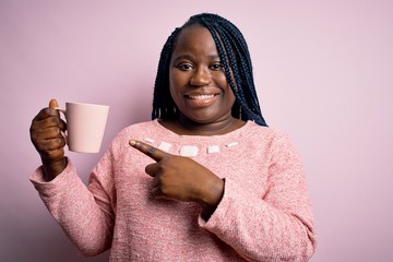 Sticker - African american plus size woman with braids drinking mug of coffee over pink background very happy pointing with hand and finger