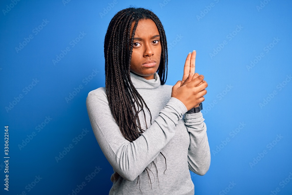 Young african american woman standing wearing casual turtleneck over blue isolated background Holding symbolic gun with hand gesture, playing killing shooting weapons, angry face
