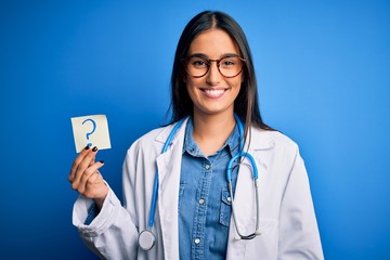 Wall Mural - Young beautiful brunette doctor woman holding paper with question mark symbol message with a happy face standing and smiling with a confident smile showing teeth