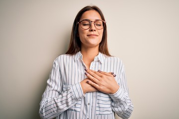 Wall Mural - Young beautiful brunette woman wearing casual shirt and glasses over white background smiling with hands on chest with closed eyes and grateful gesture on face. Health concept.