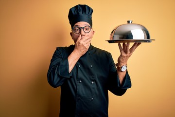 Poster - Middle age handsome grey-haired waiter man wearing cooker uniform and hat holding tray shocked covering mouth with hands for mistake. Secret concept.