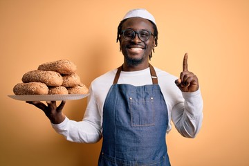 Sticker - Young african american bakery man holding tray with healthy wholemeal bread surprised with an idea or question pointing finger with happy face, number one