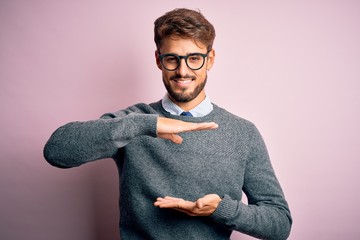 Wall Mural - Young handsome man with beard wearing glasses and sweater standing over pink background gesturing with hands showing big and large size sign, measure symbol. Smiling looking at the camera. Measuring 