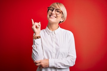 Wall Mural - Young blonde business woman with short hair wearing glasses over red background with a big smile on face, pointing with hand and finger to the side looking at the camera.