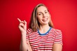 © Krakenimages.com - Young beautiful blonde woman wearing casual striped t-shirt over isolated red background with a big smile on face, pointing with hand and finger to the side looking at the camera.