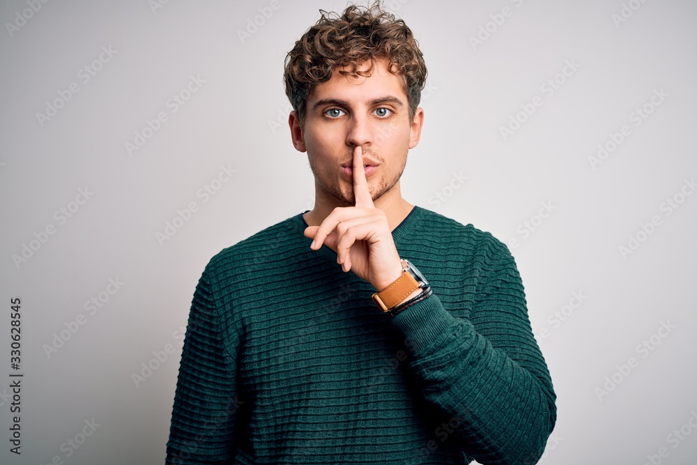 Young blond handsome man with curly hair wearing green sweater over white background asking to be quiet with finger on lips. Silence and secret concept. - obrazy, fototapety, plakaty 