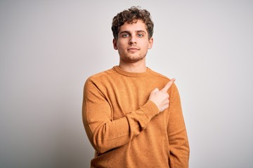 Young blond handsome man with curly hair wearing casual sweater over white background Pointing with hand finger to the side showing advertisement, serious and calm face