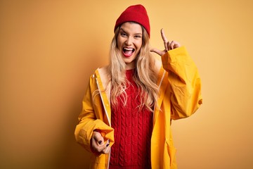 Poster - Young beautiful blonde woman wearing raincoat and wool cap over isolated yellow background pointing finger up with successful idea. Exited and happy. Number one.