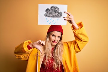 Poster - Young beautiful blonde woman wearing raincoat and wool cap holding banner with cloud image with angry face, negative sign showing dislike with thumbs down, rejection concept