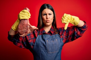 Poster - Young butcher woman holding fresh raw beef meat stake over red background with angry face, negative sign showing dislike with thumbs down, rejection concept