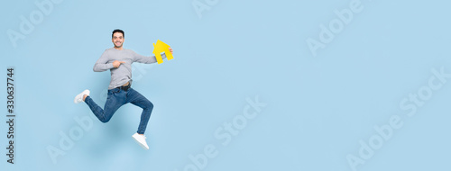 Happy handsome man jumping and pointing to yellow house model isolated on blue banner background with copy space