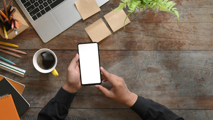 Top view image of hands holding a cropped black smartphone with white blank screen on wooden table with computer laptop, coffee cup, pencils, diary, notebook and potted plant, ruler, post it.