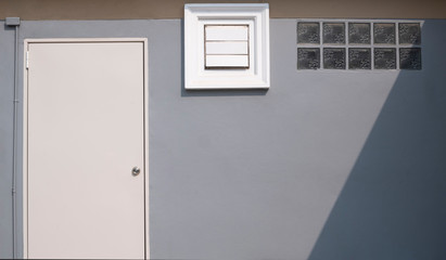 Wall Mural - Front view of white door, square ventilation shutter and glass blocks with sunlight and shade on surface of gray cement wall with brown stripe, image for home exterior decoration design concept