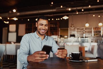 Happy african man holding mobile phone typing card data to make online payment sitting in cafe