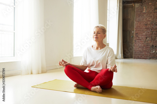 Attractive middle aged woman with short gray hair sitting on yoga mat in half lotus posture meditating after practice, keeping eyes closed and making mudra gesture, having peaceful facial expression