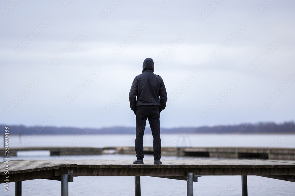 One young man in black clothes standing alone on wooden footbridge and staring at lake. Hooded guy. Peaceful atmosphere in nature. Enjoying fresh air in winter. Back view. - obrazy, fototapety, plakaty 