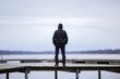 One young man in black clothes standing alone on wooden footbridge and staring at lake. Hooded guy. Peaceful atmosphere in nature. Enjoying fresh air in winter. Back view.