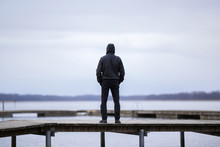 One Young Man In Black Clothes Standing Alone On Wooden Footbridge And Staring At Lake. Hooded Guy. Peaceful Atmosphere In Nature. Enjoying Fresh Air In Winter. Back View.