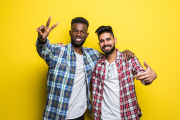 Wall Mural - Portrait of a two happy young men in sunglasses showing peace gesture over yellow background