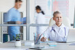 © Seventyfour - Portrait of senior female doctor using laptop while working in office with other people in background, copy space