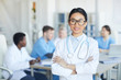 © Seventyfour - Waist up portrait of female Asian doctor standing with arms crossed and smiling at camera against medical conference background, copy space