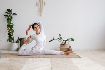 Wall Mural - Slim pretty positive young brunette woman stretching in Monkey God exercise, Splits, Hanumanasana pose on mat on floor surrounded by houseplants on white wall. Advertising space. Yoga and pilates