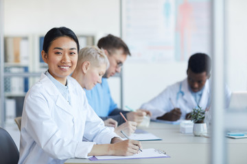 Wall Mural - Portrait of Asian female doctor smiling at camera while sitting at table during medical council or conference in clinic, copy space