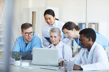 Wall Mural - multi-ethnic group of doctors using laptop together while sitting at desk in medical office interior