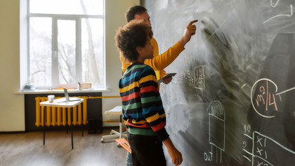 An investment in knowledge pays the best interest. Young male science teacher explaining main rules and formulas in physics and mechanics. A boy is standing near the blackboard and listening to him