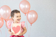 Cute Little Girl In A Pink Blouse Surrounded By Pink Balloons. The Child Holds A Congratulatory Card With A Heart. Kid Is Smiling And Happy To Receive Balloons For The Holiday