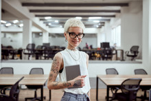 Great Working Day. Portrait Of Pretty Tattooed Blonde Businesswoman In Eyeglasses Holding Digital Tablet And Smiling At Camera While Standing In The Modern Office
