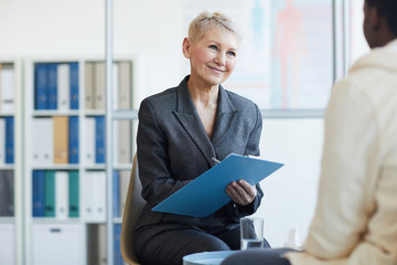 Portrait of mature female psychologist holding clipboard and smiling while consulting patient in therapy session, copy space