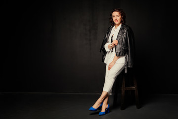 Wall Mural - Successful, happy woman in a white suit and black leather jacket sits on a chair and looks at the camera on a black background. Free space