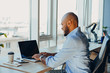 © Nana_studio - Have a nice working day. Confident young african american businessman working on laptop and talking on cell phone while sitting at his workplace in office