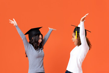 Happy excited young female students wearing graduate caps smiling with hands raising celebrating graduation day isolated on orange background