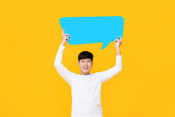 Excited smiling young cute Asian man holding blue speech bubble with empty space for text on colorful yellow background