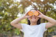 © nareekarn - photo Happy Little asian girl child standing showing front teeth with big smile. Covering eyes with orange..fresh healthy green bio background with abstract blurred foliage and bright summer sunlight.