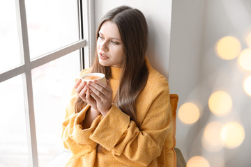 Sticker - Beautiful young woman with cup of tea sitting on window sill at home