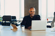 © Nana_studio - Smiling bearded african man using laptop at home, sitting on a wooden table. Concept of young people work mobile devices. Blurred window background, wide.