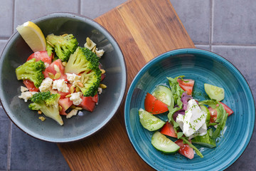 Two plates of vegetable salad with feta cheese, cucumbers, tomatoes, and broccoli served in bowl on rustic wooden board.